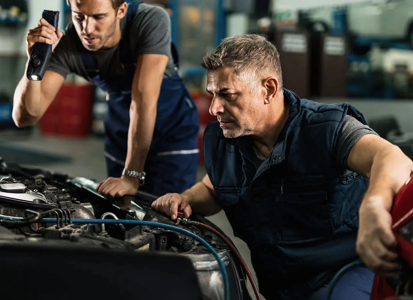 Two mechanics inspecting an open car hood in an auto repair shop, one holding a flashlight and the other looking closely at the engine components.